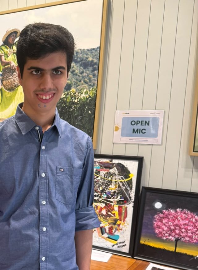 Vivaan smiling next to two of his paintings on display, a pink blossom tree and a colourful abstract